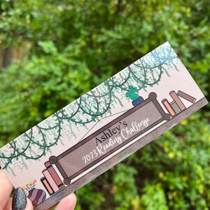 以下が含まれることがあります： 「Ashley's 2023 Reading Challenge」と書かれた看板と、つる植物が絡み付いた本棚のファンシーなイラストが描かれたしおりです。