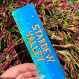 May include: A blue and gold bookmark with the text "Stardew Valley" on it. The bookmark has a pixelated design and features a small, gray bird.