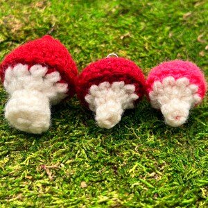 May include: Three handmade crochet mushroom ornaments. The mushroom caps are red, with white gills and stems. One mushroom is pink. The mushrooms are displayed on a bed of green moss.
