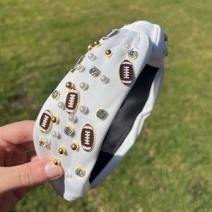 May include: White fabric headband with a knotted design, decorated with gold football charms and white pearl-like beads.
