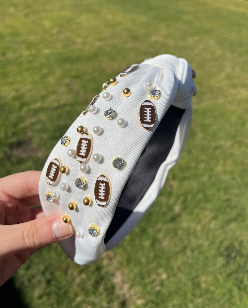 May include: White fabric headband with a knotted design, decorated with gold football charms and white pearl-like beads.
