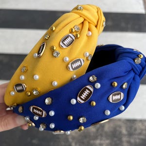 May include: Two fabric headbands with a knotted design. One headband is yellow with white pearls and gold football charms. The other headband is blue with white pearls and gold football charms.