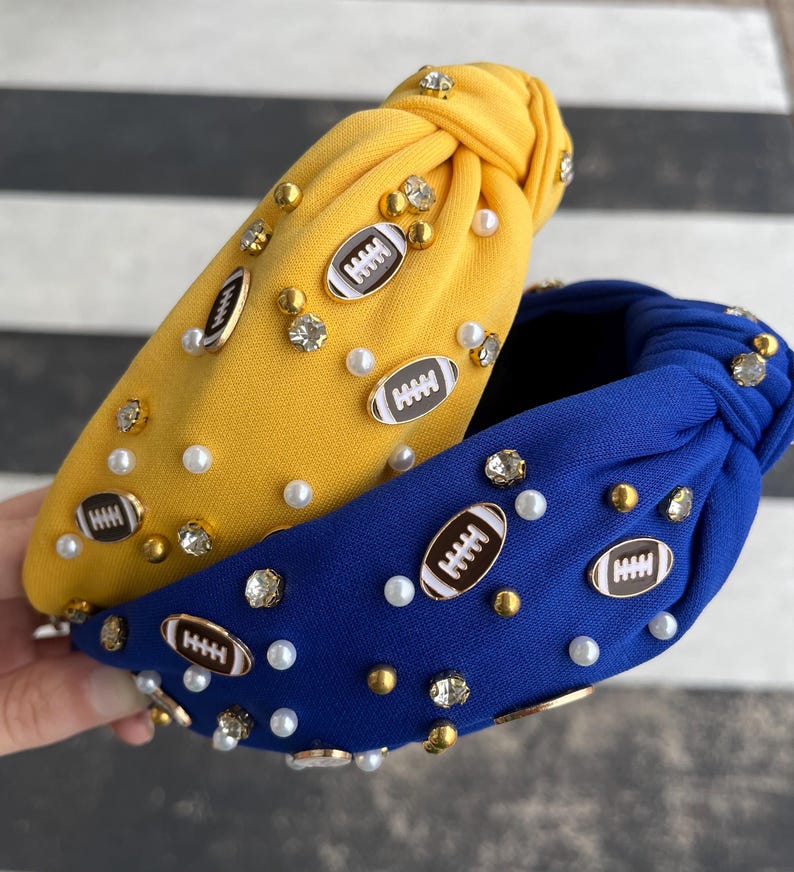 May include: Two fabric headbands with a knotted design. One headband is yellow with white pearls and gold football charms. The other headband is blue with white pearls and gold football charms.