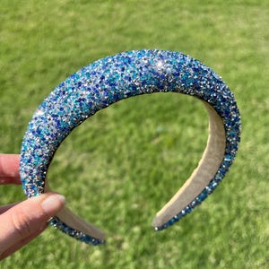 May include: A padded headband covered in sparkling blue and silver rhinestones. The headband has a light beige interior and is set against a green grassy background. The rhinestones are various shades of blue.