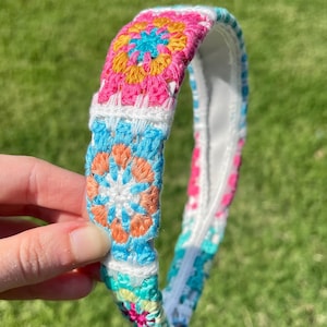 May include: A colorful crochet headband featuring a vibrant floral pattern in pink, blue, orange, and yellow. The headband is white on the inside and is held against a green grassy background.