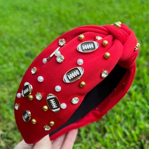 May include: Red fabric headband with a knotted design. The headband is decorated with gold and silver rhinestones, white pearls, and football-shaped charms.