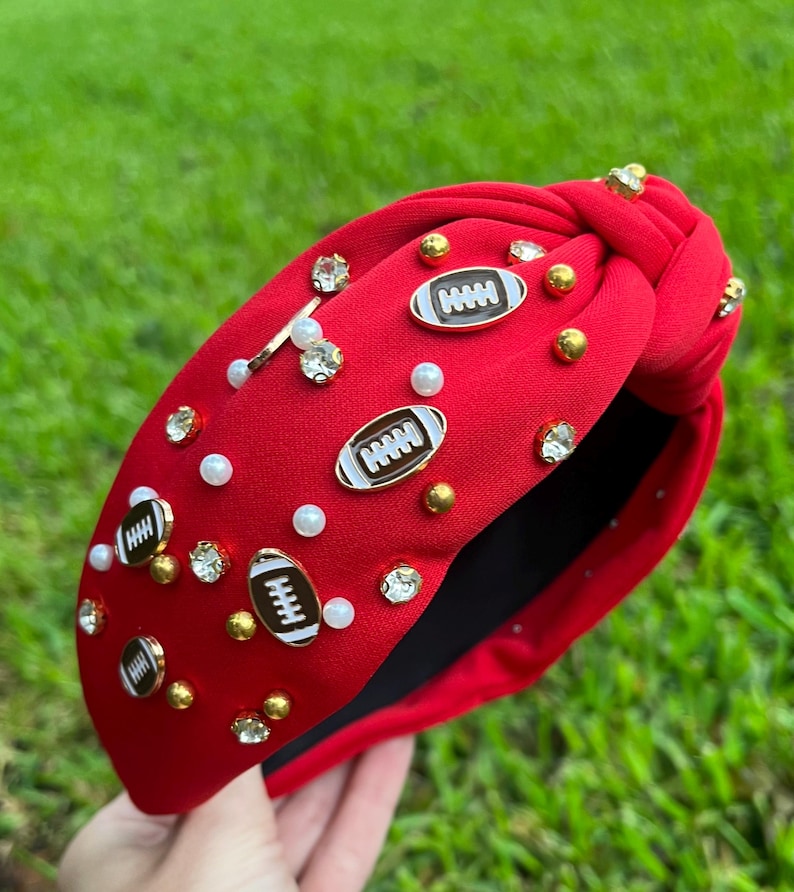 May include: Red fabric headband with a knotted design. The headband is decorated with gold and silver rhinestones, white pearls, and football-shaped charms.