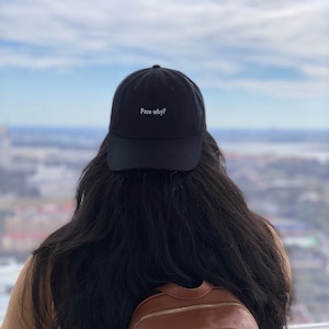 May include: A person wearing a black baseball cap with the text "Pero why?" embroidered on the front. The person has long black hair and is wearing a brown leather backpack.