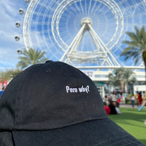 May include: A black baseball cap with white embroidery that reads "Pero why?" The cap is in focus, while a large Ferris wheel is blurred in the background.