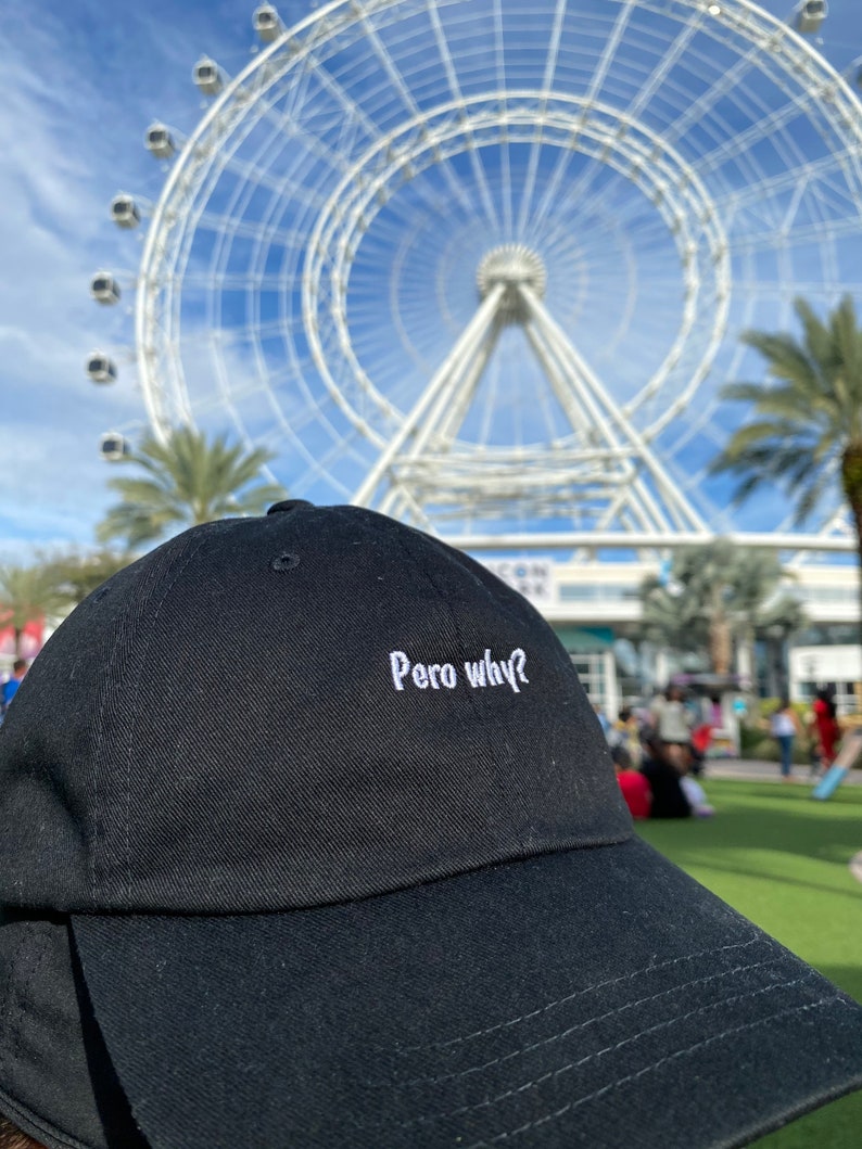 May include: A black baseball cap with white embroidery that reads "Pero why?" The cap is in focus, while a large Ferris wheel is blurred in the background.