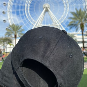 May include: A black baseball cap with a brown adjustable strap and buckle. The cap is made of a soft, textured fabric.