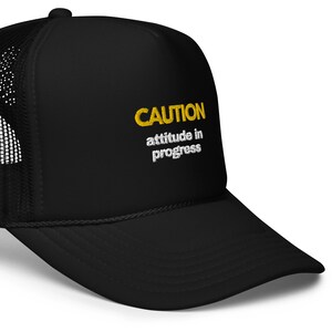 Puede incluir: Gorra de camionero negra con un mensaje bordado en amarillo que dice "CAUTION attitude in progress".