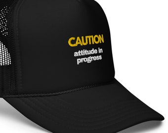 Könnte beinhalten: Schwarze Trucker-Kappe mit einer gelben Stickerei, die "CAUTION attitude in progress" lautet.