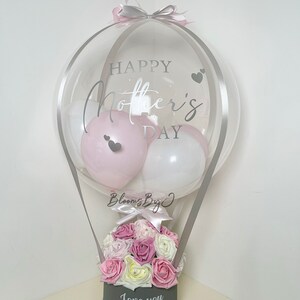 Puede incluir: Un globo transparente con texto "Happy Mother's Day" y corazones, que contiene globos rosas y blancos. Debajo, una caja gris contiene rosas rosas, blancas y moradas, con el texto "Love you Mum". Un regalo decorativo.
