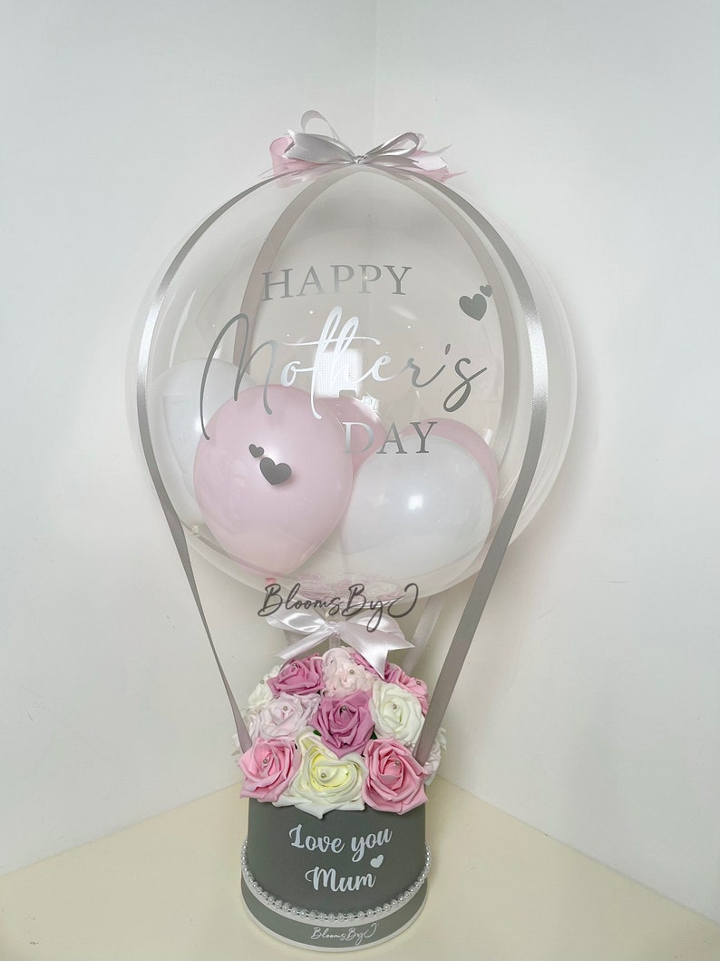 Puede incluir: Un globo transparente con texto "Happy Mother's Day" y corazones, que contiene globos rosas y blancos. Debajo, una caja gris contiene rosas rosas, blancas y moradas, con el texto "Love you Mum". Un regalo decorativo.