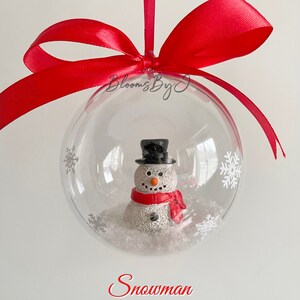 May include: A clear bauble containing a snowman figure, decorated with white snowflakes. The snowman has a black top hat and a red scarf. It is suspended by a red satin ribbon. The word "Snowman" is written in red.