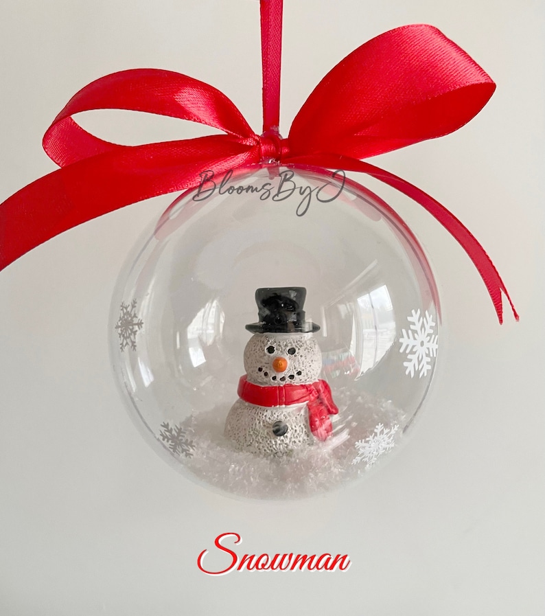 May include: A clear bauble containing a snowman figure, decorated with white snowflakes. The snowman has a black top hat and a red scarf. It is suspended by a red satin ribbon. The word "Snowman" is written in red.