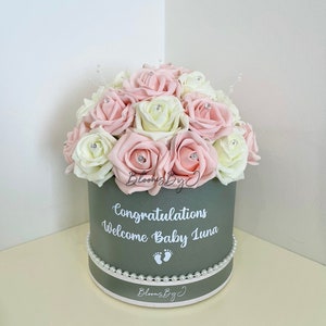 May include: A grey circular box filled with pink and cream roses, each with a gem. The box is trimmed with pearls and features the text "Congratulations Welcome Baby Luna". A baby shower gift. The box is a baby shower gift.