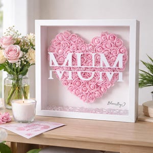 May include: A white framed floral heart with the word "MUM" in white, made of pink roses. The frame includes "Est 2016" and "BloomsByO". A lit candle and a bouquet of flowers are in the background. A greeting card is also visible.