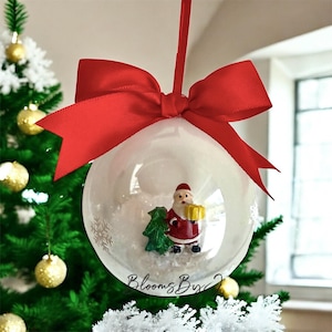 May include: A clear Christmas bauble with a miniature Santa Claus figure holding a gift and a tree inside. The bauble is suspended by a red ribbon and bow. The background includes a Christmas tree and the text "Blooms By".