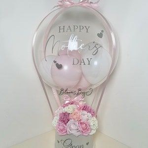 Puede incluir: Un arreglo decorativo de globo aerost&aacute;tico para el D&iacute;a de la Madre. El globo transparente tiene el texto "Happy Mother's Day" y contiene globos rosas y blancos. Un arreglo floral de rosas rosas y blancas se encuentra en una caja blanca con la palabra "Gran".