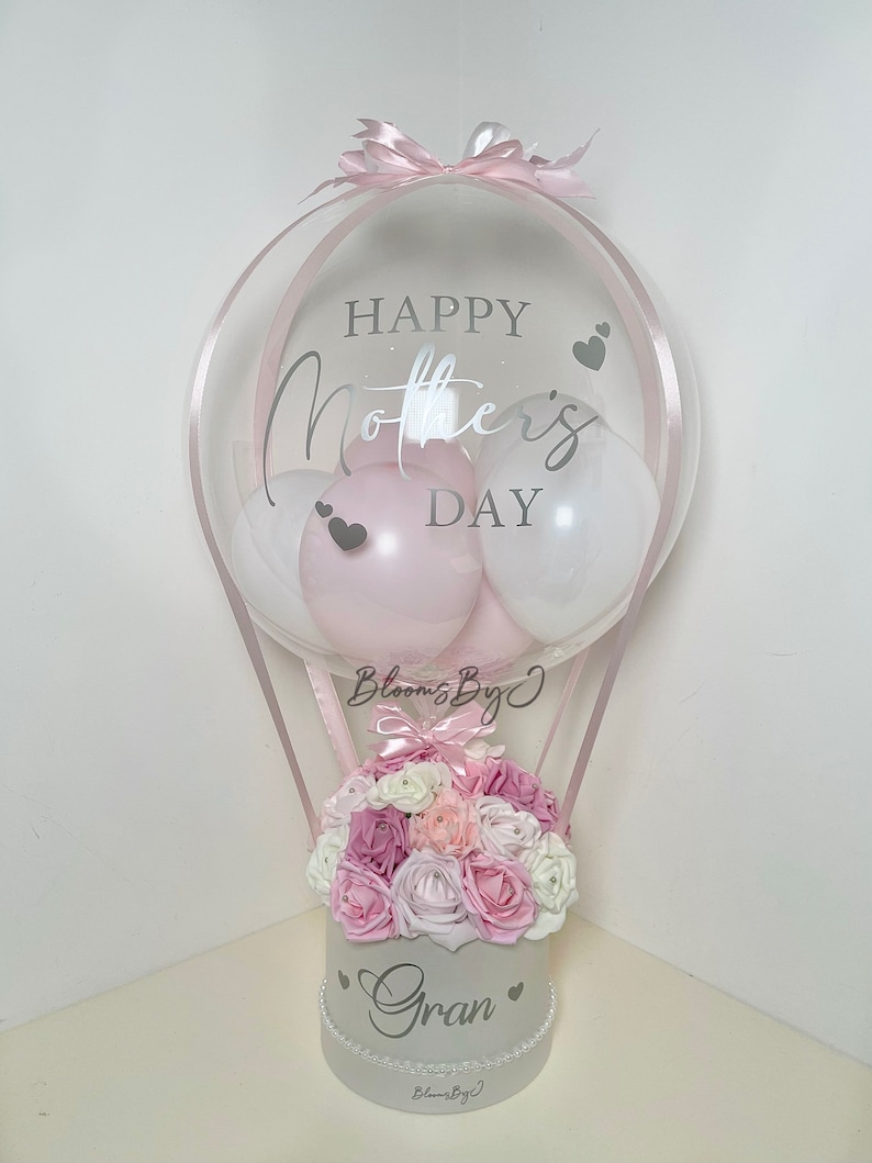Puede incluir: Un arreglo decorativo de globo aerost&aacute;tico para el D&iacute;a de la Madre. El globo transparente tiene el texto "Happy Mother's Day" y contiene globos rosas y blancos. Un arreglo floral de rosas rosas y blancas se encuentra en una caja blanca con la palabra "Gran".