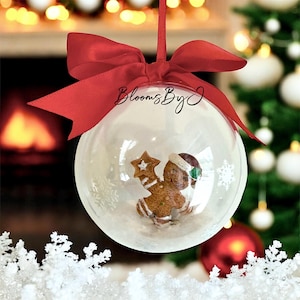 May include: A clear Christmas bauble containing a gingerbread man figurine, snowflakes, and a red ribbon. The bauble has the text "Blooms By J". The background features a Christmas tree and a fireplace.