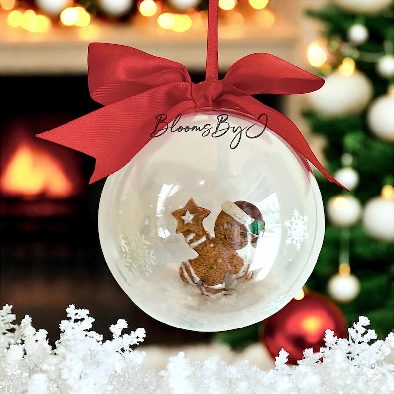 May include: A clear Christmas bauble containing a gingerbread man figurine, snowflakes, and a red ribbon. The bauble has the text "Blooms By J". The background features a Christmas tree and a fireplace.