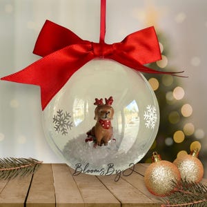 May include: A clear Christmas bauble with a brown dog figurine wearing reindeer antlers and a red scarf. The bauble has white snowflakes and is topped with a red bow. Two gold glitter baubles are on the wooden surface.