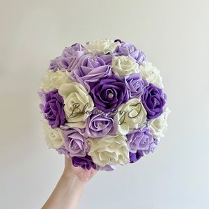 Puede incluir: Un ramo redondo de rosas artificiales en tonos morados y crema, con detalles de pedrer&iacute;a. El ramo se sostiene en una mano sobre un fondo blanco liso. Las flores est&aacute;n muy juntas, creando una forma esf&eacute;rica completa.