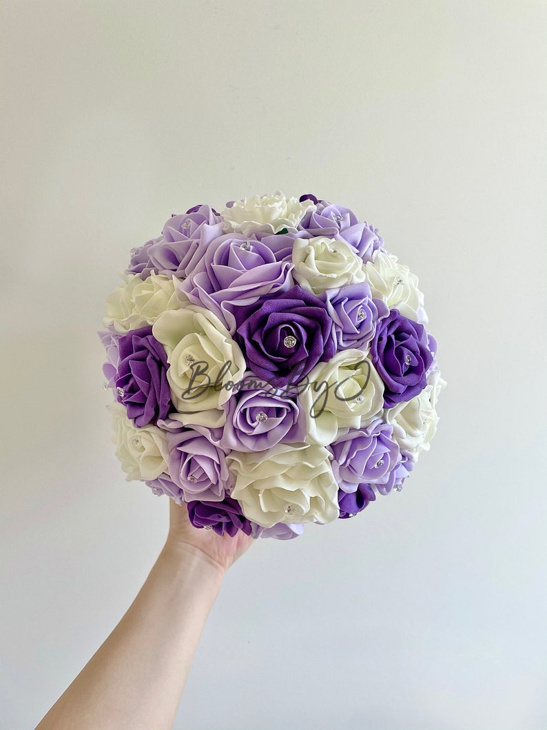 Puede incluir: Un ramo redondo de rosas artificiales en tonos morados y crema, con detalles de pedrer&iacute;a. El ramo se sostiene en una mano sobre un fondo blanco liso. Las flores est&aacute;n muy juntas, creando una forma esf&eacute;rica completa.