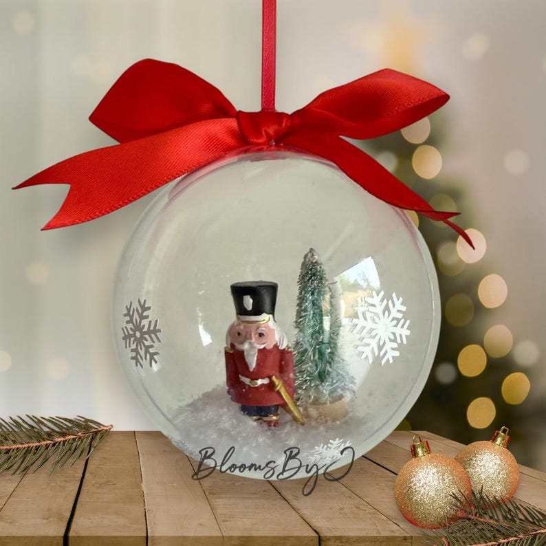 May include: A clear Christmas bauble with a red ribbon and bow. Inside the bauble is a miniature nutcracker soldier, a small Christmas tree, and white snow. The bauble has white snowflake designs.