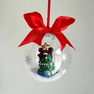 May include: A clear Christmas bauble with a green tree figurine, a gold star, and the words "Merry Christmas." The bauble is decorated with white snowflakes and a red ribbon.