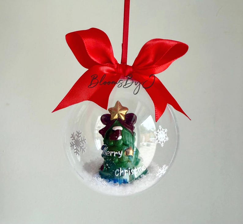 May include: A clear Christmas bauble with a green tree figurine, a gold star, and the words "Merry Christmas." The bauble is decorated with white snowflakes and a red ribbon.