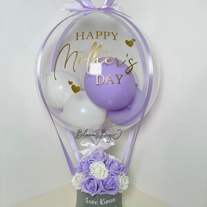 Puede incluir: Un globo transparente con la inscripci&oacute;n dorada "Happy Mother's Day", que contiene globos blancos y morados. Debajo, una caja gris contiene rosas moradas y blancas, con el texto "Love Kiran and Jasmine".