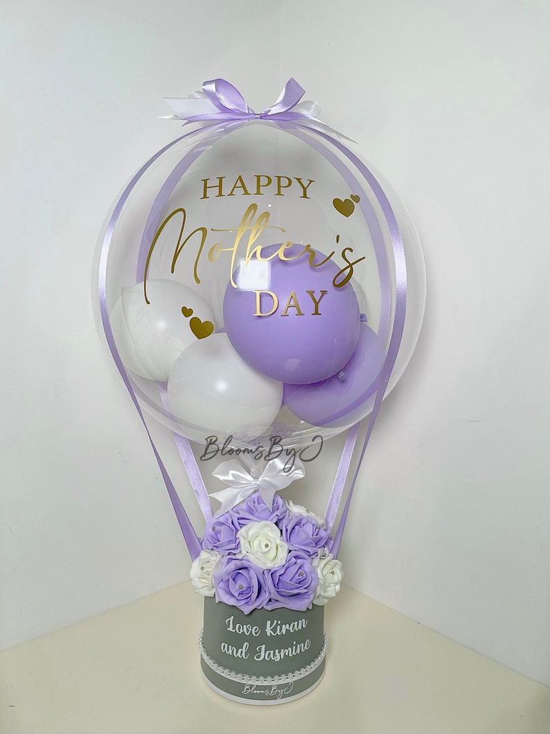 Puede incluir: Un globo transparente con la inscripci&oacute;n dorada "Happy Mother's Day", que contiene globos blancos y morados. Debajo, una caja gris contiene rosas moradas y blancas, con el texto "Love Kiran and Jasmine".