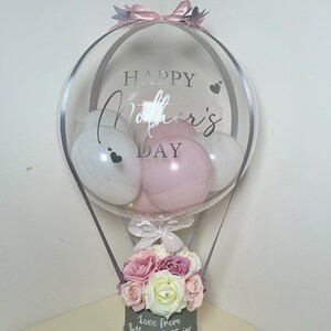Puede incluir: Un arreglo de globos decorativo para el D&iacute;a de la Madre. El globo transparente con el texto "HAPPY Mother's DAY" contiene globos blancos y rosas. La base es una caja gris llena de flores rosas y blancas, con el texto "Love from Beth and Claire".