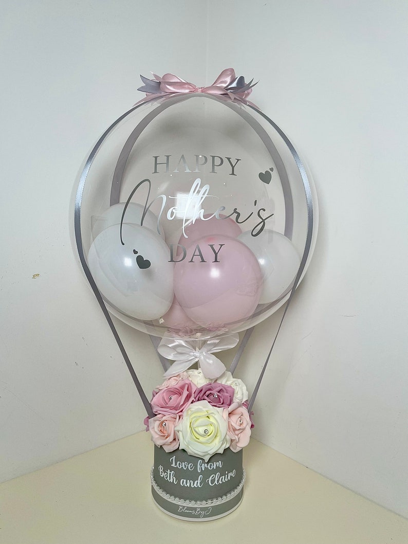 Puede incluir: Un arreglo de globos decorativo para el D&iacute;a de la Madre. El globo transparente con el texto "HAPPY Mother's DAY" contiene globos blancos y rosas. La base es una caja gris llena de flores rosas y blancas, con el texto "Love from Beth and Claire".