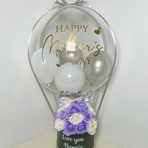 Puede incluir: Arreglo decorativo de globo aerost&aacute;tico para el D&iacute;a de la Madre. El globo transparente est&aacute; lleno de globos blancos y plateados y presenta letras doradas que dicen "Happy Mother's Day". La base es una caja verde oscuro con flores moradas y blancas y el texto "Love you Nannie."