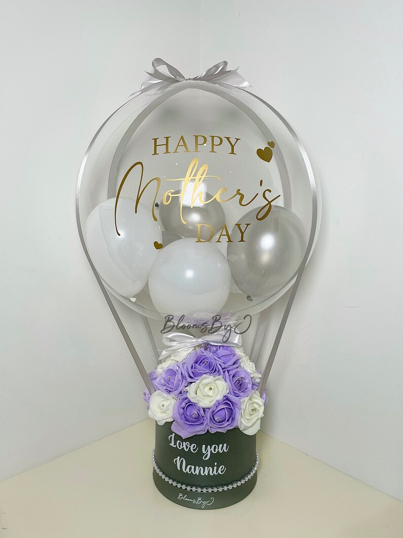 Puede incluir: Arreglo decorativo de globo aerost&aacute;tico para el D&iacute;a de la Madre. El globo transparente est&aacute; lleno de globos blancos y plateados y presenta letras doradas que dicen "Happy Mother's Day". La base es una caja verde oscuro con flores moradas y blancas y el texto "Love you Nannie."