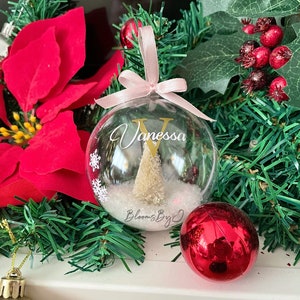 May include: A clear Christmas bauble with the name "Vanessa" and a gold initial "V". Inside the bauble is a small Christmas tree and faux snow. The bauble hangs from a pink ribbon. Red and silver baubles and a red poinsettia are in the background.