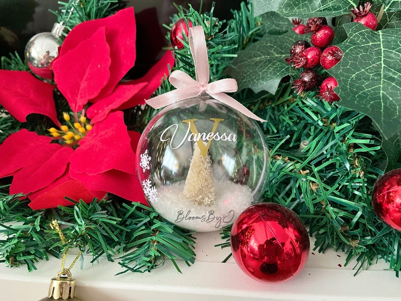 May include: A clear Christmas bauble with the name "Vanessa" and a gold initial "V". Inside the bauble is a small Christmas tree and faux snow. The bauble hangs from a pink ribbon. Red and silver baubles and a red poinsettia are in the background.