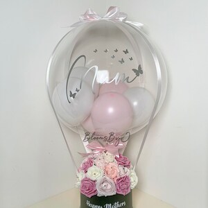 Puede incluir: Un globo transparente con la palabra "Mam" y calcoman&iacute;as de mariposas, que contiene globos blancos y rosas. Debajo, una caja negra muestra "Happy Mothers Day" y est&aacute; llena de flores rosas y blancas. Un lazo rosa corona el globo.