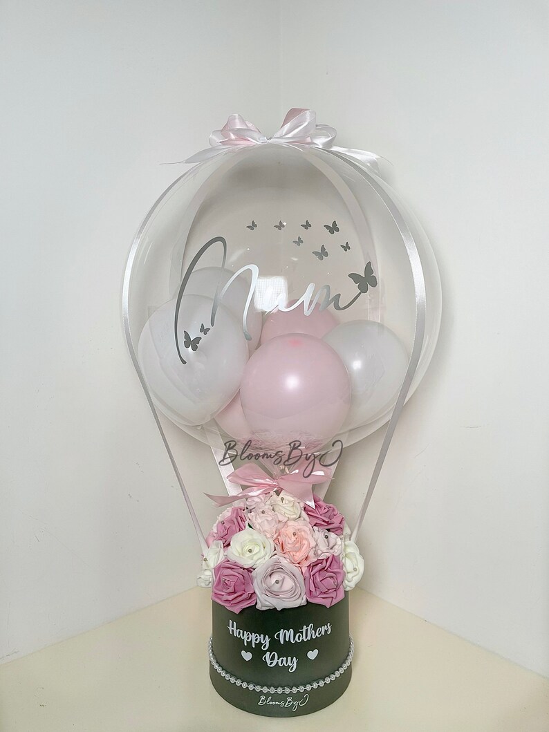 Puede incluir: Un globo transparente con la palabra "Mam" y calcoman&iacute;as de mariposas, que contiene globos blancos y rosas. Debajo, una caja negra muestra "Happy Mothers Day" y est&aacute; llena de flores rosas y blancas. Un lazo rosa corona el globo.