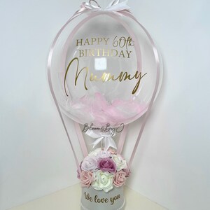 Puede incluir: Arreglo decorativo en forma de globo aerost&aacute;tico. Un globo transparente con plumas rosas y texto dorado que dice "Happy 60th Birthday Mummy" se encuentra sobre una caja gris llena de flores. La caja tiene el texto "We love you".