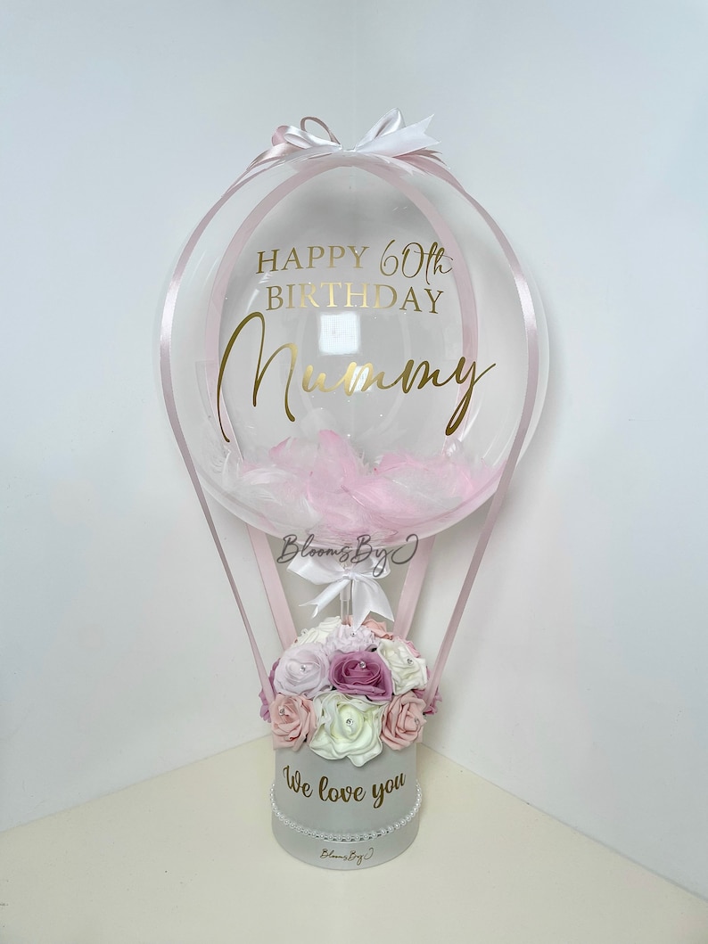 Puede incluir: Arreglo decorativo en forma de globo aerost&aacute;tico. Un globo transparente con plumas rosas y texto dorado que dice "Happy 60th Birthday Mummy" se encuentra sobre una caja gris llena de flores. La caja tiene el texto "We love you".