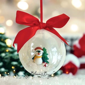 May include: A clear Christmas bauble with a red ribbon bow. Inside the bauble is a penguin wearing a Santa hat and scarf, a small Christmas tree, and a wrapped gift. The bauble has the text "Blooms By" on the bottom.