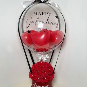 May include: A Valentine's Day gift arrangement in the shape of a hot air balloon. The clear balloon is decorated with the words "Happy Valentines Day" and contains heart-shaped red balloons. The base is a red box filled with red roses and the words "love you".