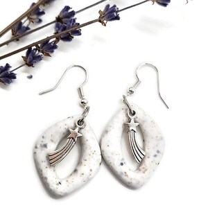 May include: A pair of white speckled earrings with a silver shooting star charm. The earrings are teardrop-shaped and hang from silver hooks. Lavender sprigs are in the background.