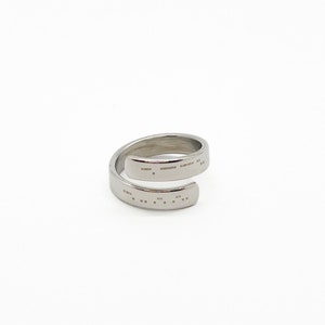 May include: A silver-colored wrap ring with a Morse code design. The ring has a spiral shape and features a series of small, rectangular markings. The ring is set against a white background.